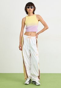 Multikleurige cropped tanktop (gele, paarse gradient) en witte trainingsbroek met zijstrepen en oranje accenten. Model draagt zonnebril.