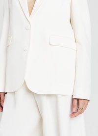 Blazer blanc ajusté avec un col cranté, deux boutons ronds et des poches latérales. Le tissu semble lisse avec une coupe structurée.