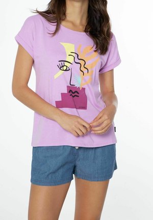 Femme portant un t-shirt lavande avec un graphisme abstrait de visage et un short en jean bleu, les mains jointes devant, sur un fond blanc.