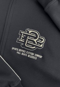 Zwarte sportkleding met een geborduurd logo in crème, met de tekst "SPORTS INSPIRED CLOTHING COMPANY" eronder. Gestructureerde stof, atletisch ontwerp.