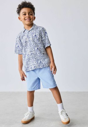 Smilende dreng stående med spredte fødder, iført en hvid kortærmet skjorte med blåt labyrintmønster, lyseblå shorts, hvide sokker og sneakers.