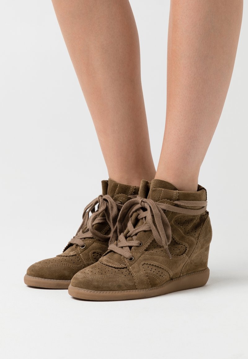 Baskets compensées en daim marron avec lacets, portées sur des jambes nues sur un fond blanc uni.