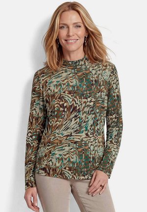 Vrouw met schouderlang blond haar, die een hoogsluitende, lange mouwen patroon top in aardetinten en beige broek draagt, glimlacht zachtjes.