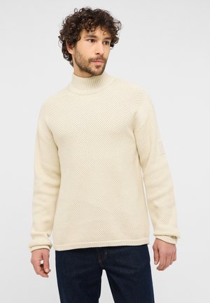 Cream-colored knitted sweater with a high collar, textured pattern, and ribbed cuffs. Features a small logo patch on the left sleeve.