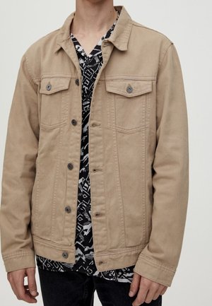 Homme portant une veste en jean beige boutonnée par-dessus une chemise à motifs noir et blanc, avec un pantalon noir, debout devant un fond uni.