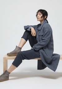 Abrigo de lana gris con botones, llevado sobre una camisa azul clara. El modelo muestra pantalones cortos azul marino y zapatillas altas grises en un banco de madera.