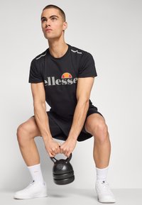 Melns sporta krekls ar pelēkiem akcentiem un logo, melni šorti, balti skriešanas apavi un melns kettlebells, kas tiek pacelts.