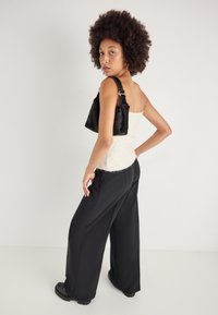 Bolsa preta em pele sintética com uma forma estruturada, usada sobre um top de malha branco. O modelo usa calças largas pretas e botas pretas robustas.