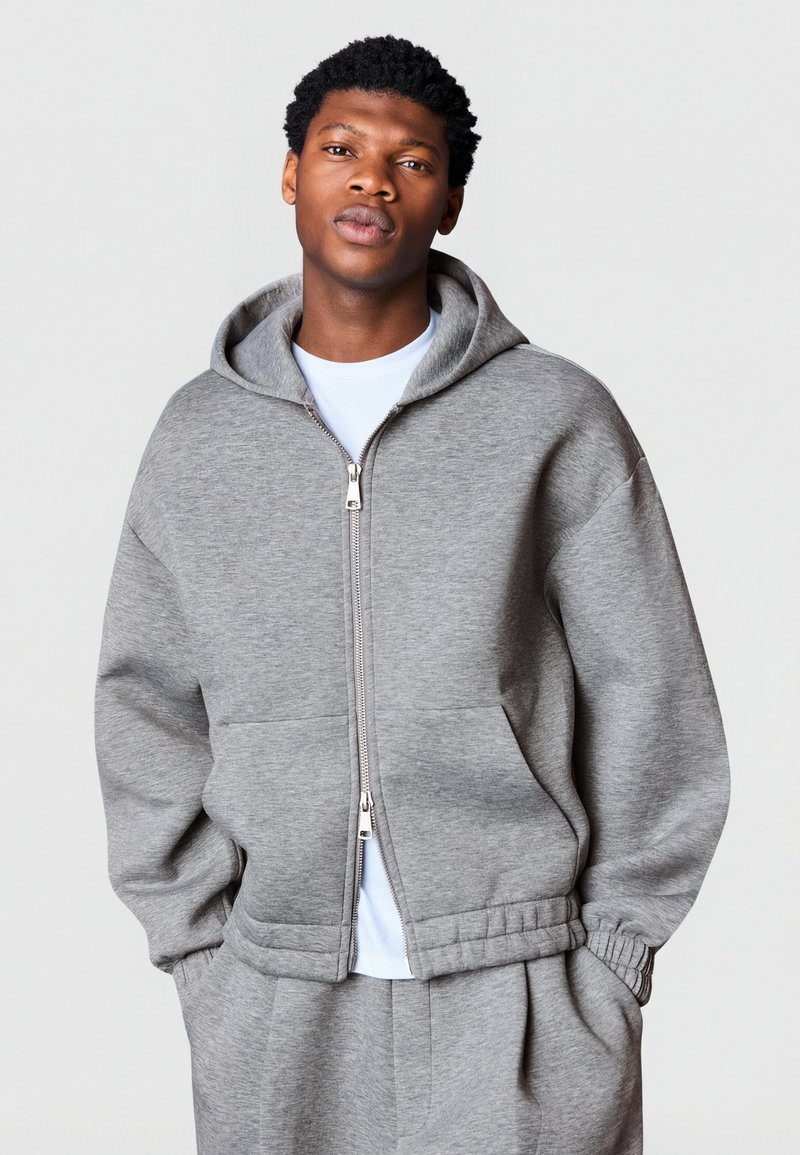 Junger Mann trägt einen grauen Kapuzenpullover mit Reißverschluss und passende Jogginghose, steht mit den Händen in den Taschen vor einem einfarbigen Hintergrund.