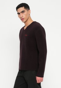 Σκούρο μπορντό πουλόβερ με v λαιμόκοψη σε μαλακό, υφασμάτινο υλικό με διακριτική ετικέτα λογότυπου. Διαθέτει μακριά μανίκια και χαλαρή εφαρμογή.