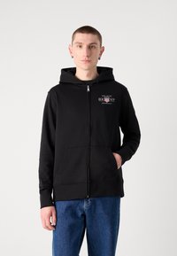 Crna zip majica s kapuljačom i prednjim džepom, materijal od pamuka, s malim logom na prsima. Nosena preko plavih traperica. Jednostavan, ležeran dizajn.