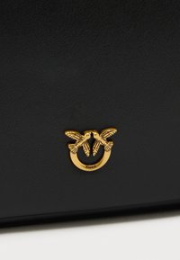 Μαύρη δερμάτινη τσάντα με χρυσό μεταλλικό διακοσμητικό σε σχήμα πουλιού και κυκλικό έμβλημα. Υφή με ελάχιστο σχεδιασμό.