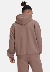 Felpa con cappuccio marrone dal taglio comodo, maniche e orlo a coste, morbida al tatto e con tasca a marsupio. Indossata con pantaloni da jogging coordinati.