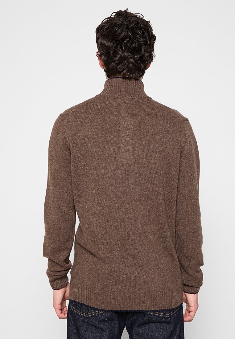 Uomo con capelli ricci e scuri, in piedi di spalle, che indossa un maglione a collo alto marrone e jeans blu scuro, su uno sfondo semplice.