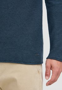 Dziewiarski granatowy sweter o ryfrowanej fakturze, z metką. Rękaw sięga do nadgarstka, zestawiony z jasnymi spodniami.