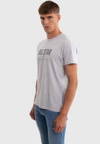 Férfi világosszürke "BIG STAR" logós pólóban és kék farmerben, homogén világosszürke háttér előtt állva.