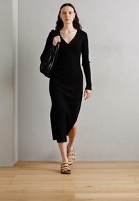 Vestido de malha preto com mangas longas, decote em V e abertura lateral. Combinado com sandálias de plataforma beje e uma mala de ombro texturizada preta.