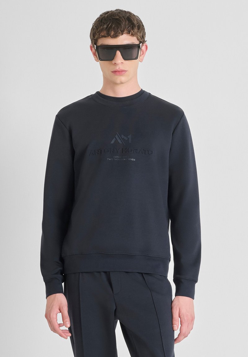 Sudadera marina con cuello redondo, que presenta un diseño de logotipo texturizado en un tono más oscuro. Combinada con pantalones a juego.