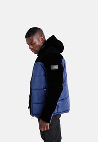 Giubbotto imbottito con corpo trapuntato blu navy e maniche in velluto nero, dotato di chiusura con zip e patch del logo sulla manica.