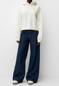 Felpa crema con tasca a marsupio frontale e orlo a coste, abbinata a jeans in denim blu scuro a gamba larga. Sneakers bianche con suola beige.