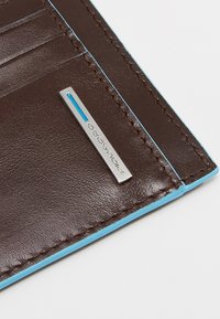 Cartera de cuero marrón con costuras y ribete azul, que cuenta con múltiples ranuras para tarjetas y una placa de logo plateada. Textura suave con líneas limpias.