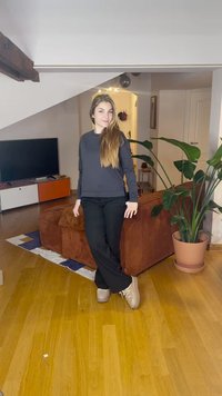Pantaloni neri larghi e un top grigio a maniche lunghe. L'ambiente presenta un pavimento in legno, un divano marrone con trama e una pianta verde in vaso.