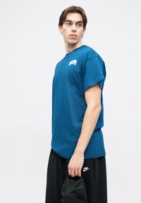 Μπλε βαμβακερό T-shirt με κοντά μανίκια, στρογγυλή λαιμόκοψη και γραφικό μπροστά. Συνδυασμένο με μαύρες βερμούδες, κρατώντας ένα μαύρο καπέλο στο χέρι.