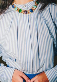 Blusa a righe blu e bianche con pieghe, caratterizzata da una collana statement dorata con pietre verdi, blu e viola. Dettaglio con bottoni sul colletto.