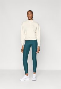 Sweat-shirt à fermeture éclair beige avec un logo sur la poitrine et des leggings teal. Modèle portant des baskets blanches, se tenant devant un fond minimaliste.