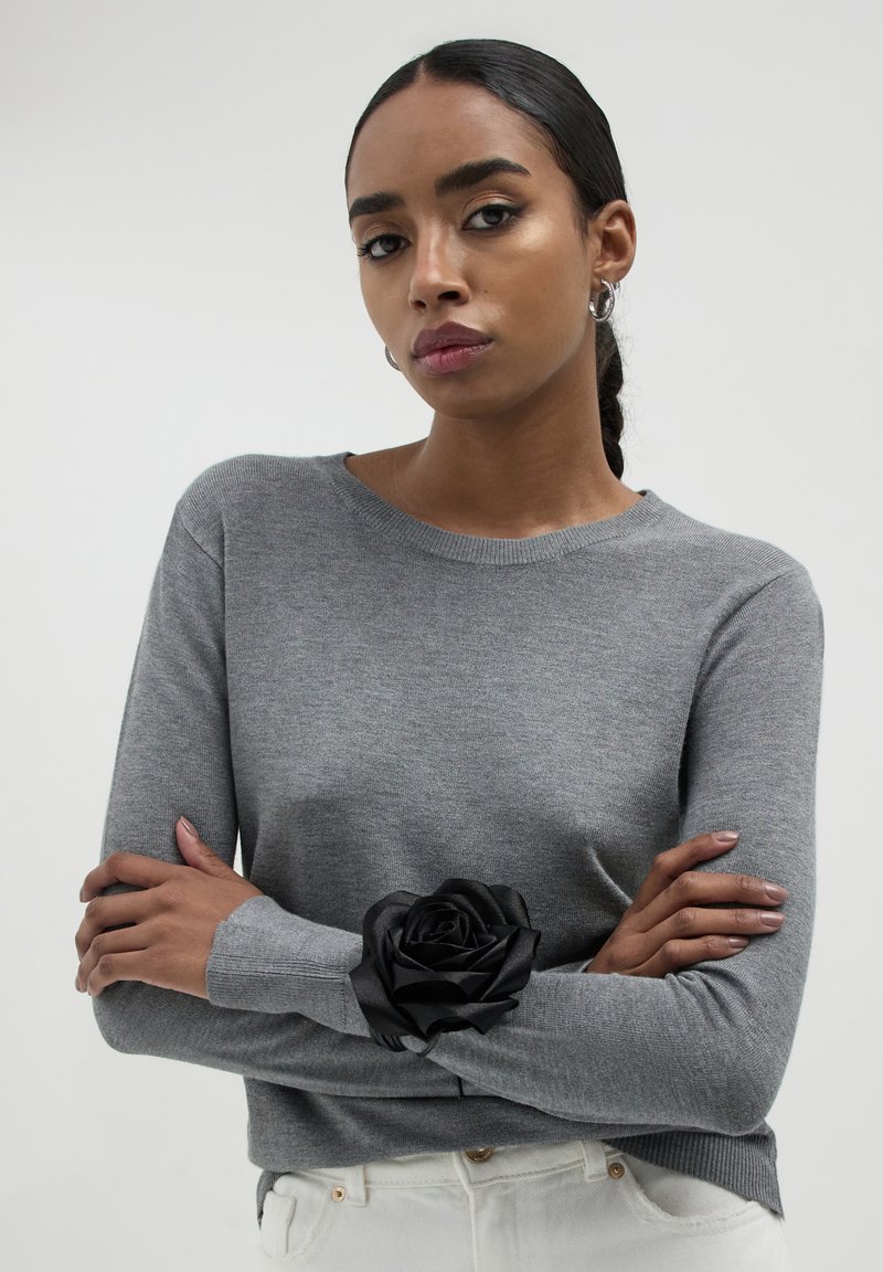 Grauer Strickpullover mit rundem Ausschnitt und langen Ärmeln, verziert mit einer großen schwarzen Satinrose auf dem linken Ärmel.
