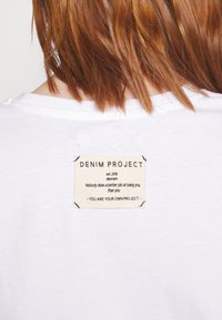 Valkoinen puuvillapaita, jossa on ommeltu suorakulmainen etiketti mustalla tekstillä beigellä taustalla, lukien "DENIM PROJECT" ja lisätietoja.