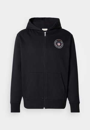 Black zip-up hoodie with front pockets, a hood, and a circular GANT logo with stars and stripes on the left chest.
