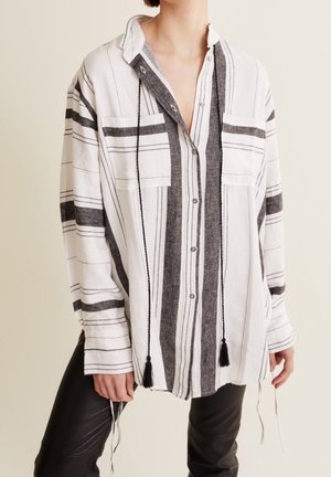 Personne portant une chemise blanche ample à boutons avec des rayures verticales et horizontales noires, deux poches poitrine et des glands noirs, associée à un pantalon noir.