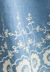 Modrá látka s propracovanou bílou květinovou výšivkou, zahrnující vinoucí se vnější prvky a kulaté motivy s jemnými perforacemi.