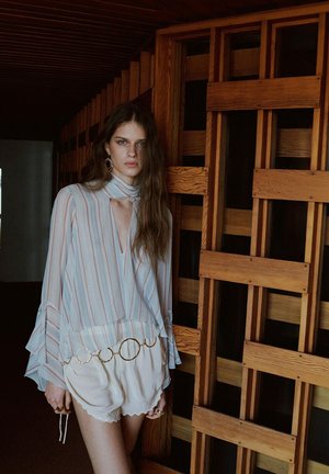 Jeune femme aux longs cheveux portant un chemisier rayé à col ouvert, un short blanc et une ceinture avec anneau doré, debout près d'un mur en treillis en bois à l'intérieur.