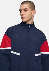Giacca a vento navy con accenti rossi e bianchi, collo alto, chiusura con zip e tessuto testurizzato. Presenta un dettaglio del logo sul petto.