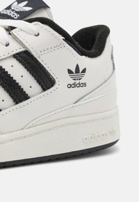 Fehér sneaker fekete részletekkel, három csíkkal, texturált bőrből készült és gumitalppal. Az Adidas logó a cipő oldalán és a nyelvén látható.