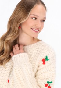 Krémový pletený sveter s veľkými stehnami, s motívmi červených čerešní na ramenách. Modelka má dlhé, vlnité vlasy a náušnice v tvare červených čerešní.