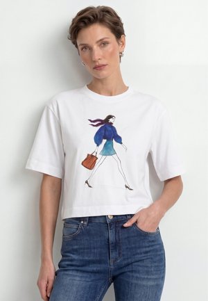 Frau mit kurzen Haaren, die ein weißes T-Shirt mit der Illustration einer gehenden Frau in blauem Outfit und brauner Tasche trägt, kombiniert mit blauen Jeans.