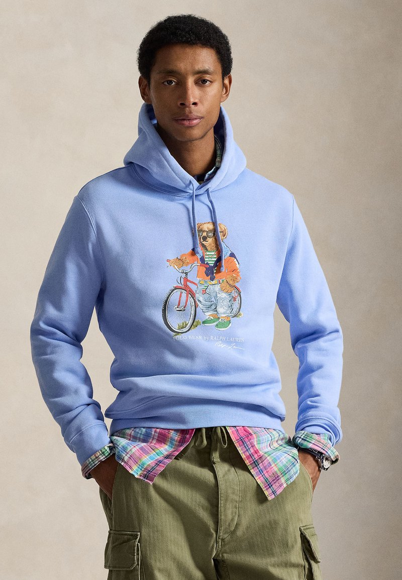 Ανοιχτό μπλε φούτερ με κουκούλα, με εικόνα αρκούδας πάνω σε ποδήλατο. Συνδυασμένο με πουκάμισο καρό. Παντελόνι καρό με τσέπες. Καθημερινό στυλ δρόμου.