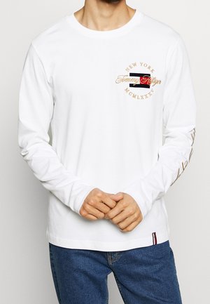 Weißes Langarmshirt mit einem runden Logo in Gold und Rot auf der Brust, mit Text und gestreiften Akzenten nahe dem Saum.