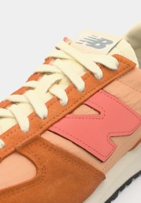 Baskets avec un dessus en daim orange et en tissu pêche, lacets blancs, grand logo "N" rose, et un logo gris sur la languette. Semelle en caoutchouc noir.