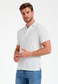 Camisa polo blanca con collar, mangas cortas, tapeta de dos botones y aberturas laterales. Tejido suave con un diseño entallado y un detalle sutil del logo.