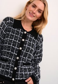 Melnā un baltā ruļļotā krāsā adīta kardigāna ar trikotāžas šuvu, lieliem pērļu pogām un melnu apkakli. Satur sānu kabatas un brīvu piegriezumu.