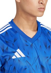 Blaues Sporttrikot mit geometrischem Muster, V-Ausschnitt und weißem Adidas-Logo sowie drei weißen Streifen auf den Schultern.
