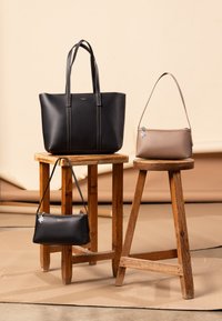 Drie tassen weergegeven op houten krukken: een grote zwarte tote, een middelgrote zwarte handtas en een kleine beige crossbody. Allemaal gemaakt van glad leer met subtiele branding.