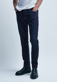 Mörkblå denimjeans med slim fit och subtil textur, tillsammans med svarta läderchelsea-boots. Inga synliga dekorationer.