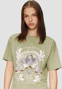 Oliwkowa bawełniana koszulka z dużym graficznym nadrukiem stylizowanych kwiatów i geometrycznych kształtów w stonowanej purpurze i bieli.