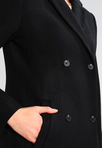 Sort dobbeltradet frakke lavet af blødt stof, med store sorte knapper, en spidse krave og sidelommer.