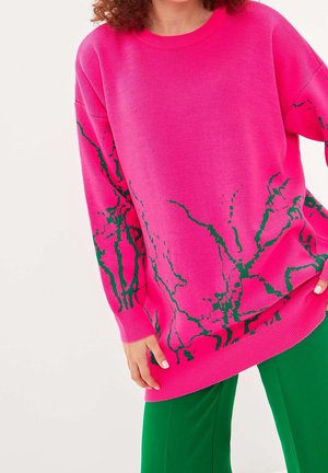Personne portant un pull oversize rose vif avec des motifs abstraits verts et un pantalon vert, debout devant un fond uni.